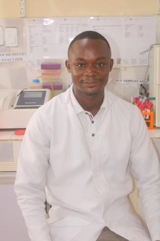 A man in a white lab coat smiling at the camera in a laboratory setting with equipment and supplies visible in the background.
