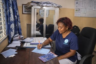 Woman in navy scrubs writing or reviewing documents at a desk in a medical office with a window and filing cabinet visible in the background.
