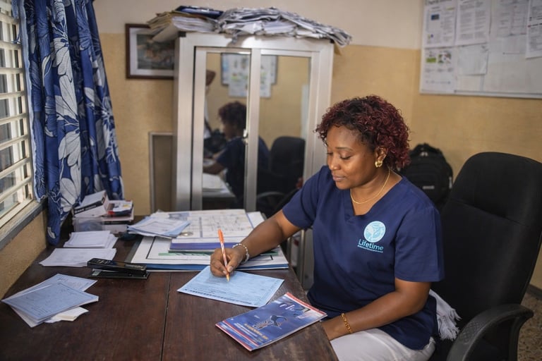 Woman in navy scrubs writing at desk in medical office, surrounded by papers and documents