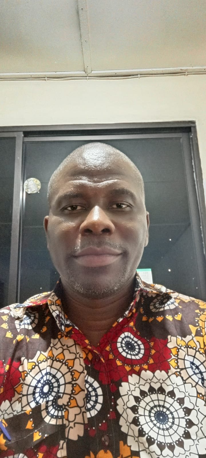 Self-portrait of a man with a shaved head wearing a colorful patterned shirt with geometric designs in front of a dark door